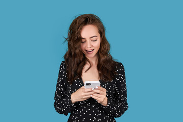 Fototapeta premium Portrait of a beautiful girl on a blue background, a girl with a phone in her hands, looks at the phone 