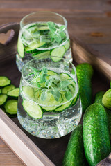 Detox drink. Water with pieces of cucumber, ice and mint leaves in transparent glasses and vegetables on a wooden tray on the table. Vertical view