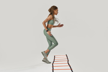 Fototapeta na wymiar Life is movement. Full length shot of young sportive mixed race woman in sportswear training on agility ladder drill isolated over grey background. Fitness workout concept