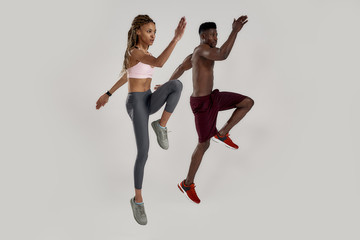 Fototapeta na wymiar Full length shot of young muscular african american man and sportive mixed race woman looking focused while jumping, exercising isolated over grey background. Sports and workout concept