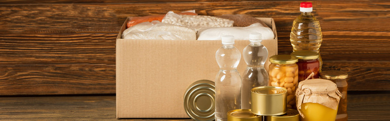 panoramic shot of cardboard box with groats near water, oil, canned food and honey on wooden background, charity concept