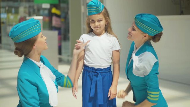 Portrait of cute little Caucasian girl smiling at camera as stewardess putting hat on her head. Happy satisfied child waiting for departure with accompanying professional flight attendants.