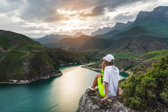 A beautiful girl tourist sits on a stone high in the mountains admiring the lake and sunset. The concept of tourism, travel and outdoor recreation