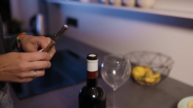 Young woman's hands with LGBT rainbow bracelet use corkscrew to open bottle of red wine in kitchen close view slow motion