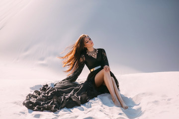 Mysterious woman in a black long dress sits in desert. Luxurious clothes, gold accessories, pretty...