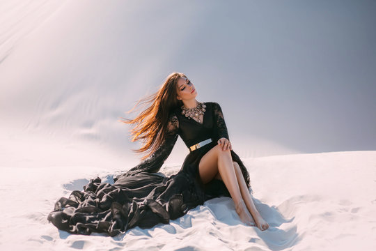 Mysterious woman in a black long dress sits in desert. Luxurious clothes, gold accessories, pretty face. Oriental fashion model. Sand dunes background. Art photo. Happy girl with flying long hair