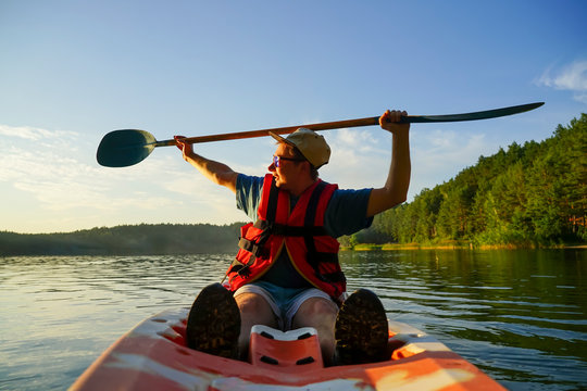 A Young Guy In A Campground And Glasses Sits In A Kayak And Holds An Oar Over His Head. On The Sparing Plan, A Lake And A Coniferous Forest