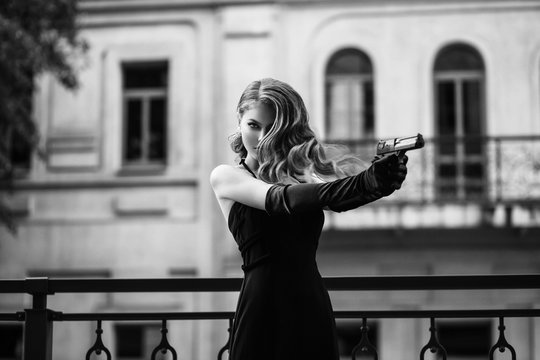 Beautiful young girl in an elegant dress. Woman holding a gun in her hands. Fashion model in the form of a dangerous sexy spy. Black and white photography. Retro lady, vintage hairstyle evening makeup
