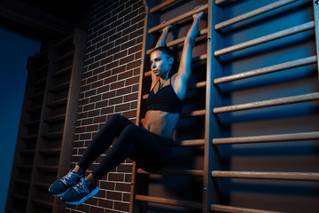 Confident fit young brunette woman doind exercises for abs on the wall bars in gym in neon lights.