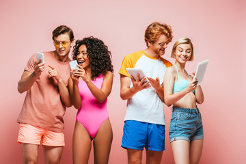 young multicultural friends in summer outfit using digital devices on pink