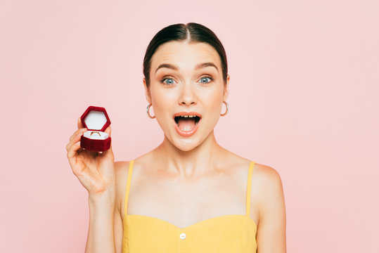 Shocked Brunette Young Woman Holding Box With Engagement Ring Isolated On Pink