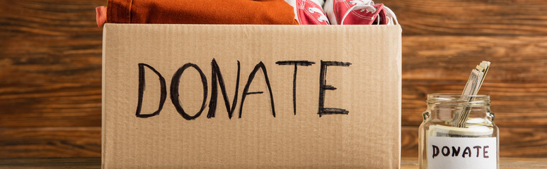panoramic shot of money in jar near cardboard box with donate lettering and clothes on wooden background, charity concept