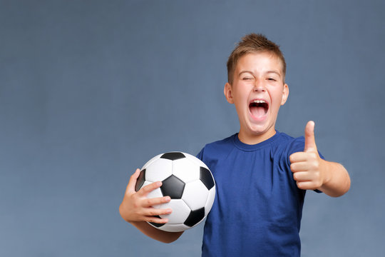 Handsome laughing fan boy holds soccer ball and gesturing thumbs up on gray background. Copy space for text. 