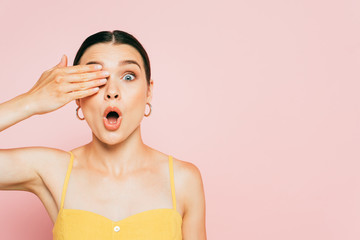 surprised brunette young woman covering eye isolated on pink