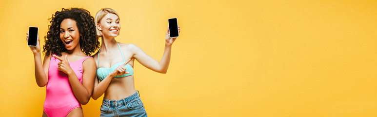 website header of young interracial women in summer outfit pointing at smartphones with blank screen isolated on yellow