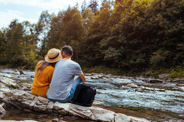 Couple of tourists relaxing by mountain river enjoying landscape. Travelers sitting on rock. Summer...