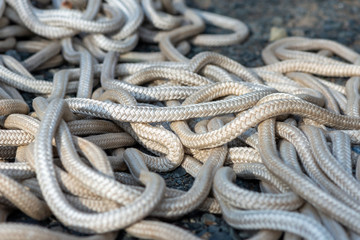 A cluster of white hallow double braided nylon commercial fishing rope piled and tangled. The rope has a 2 inch diameter and is laying on and asphalt ground. The marine rope is slightly worn and used.