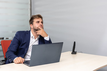 Thoughtful young business man in office sitting at work desk in front of laptop