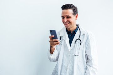 Black haired doctor wearing white coat and stethoscope looking at his mobile phone and smiling while making a video call, on white background. Medicine concept