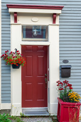 A red exterior door of a building with white and red trim on a light blue clapboard wall. The house has an antique glass window over the door, black mailbox, flowers and a decorative molding,  