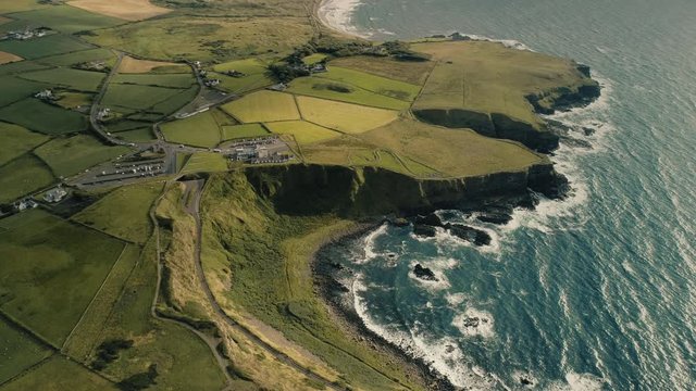 Ireland farms buildings aerial: cottages, houses with rural road at greenery meadows and yellow fields. Wonderful Irish autumn landscape at rocky shore of blue sea bay. Footage shot in 4K, UHD