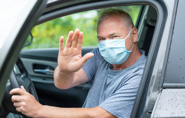 A man with a medical mask sitting behind the wheel of a car makes a stop gesture with his hand
