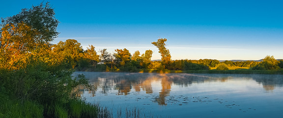 Morning on the river coast, trees reflected in the crystal clear water, on the river above the water fog.