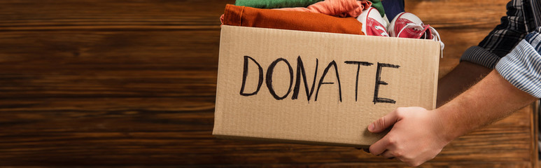 cropped view of man holding cardboard box with donate lettering and clothes on wooden background,...
