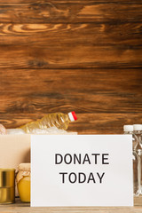 cardboard box with food and donate today card on wooden background, charity concept
