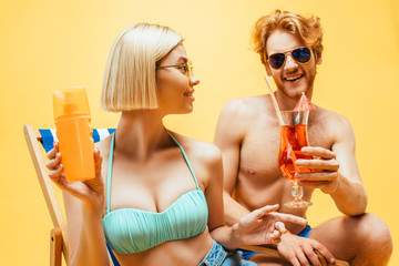 blonde woman with orange juice and young man with cocktail glass looking at each other isolated on yellow