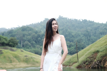 Smiling beautiful young Asian girl wear white dress standing on grass meadow beside the stream in tropical rainforest.