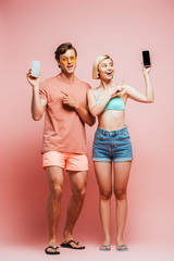 Young couple in flip flops and sunglasses pointing with fingers at smartphones on pink background