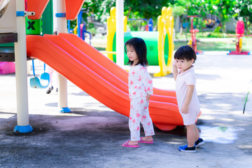 Asian sisters and brothers are choosing rides on playground. Daughter and son look at something from inside of playground. Background is orange slides and other colorful rides. Children 2-3 years old.