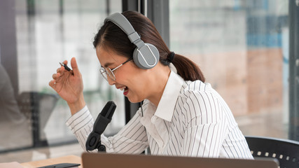 Asian women podcaster podcasting and recording online talk show at studio using headphones,...