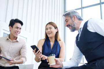 Smiling Asian team and Senior Caucasian businesspeople teamwork relaxing and brainstorm. Businesswoman holding smartphone and talking together at office. Focus asian woman.