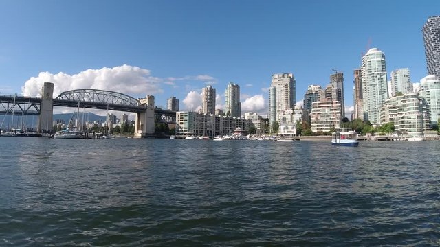 sunny slow smooth panaramic Granville Island dock facing across quayside the harbor to Yaletown Condominiums Burrard Bridge Vancity Lofts as water taxis bus drive by and some yatchs sail boats docked