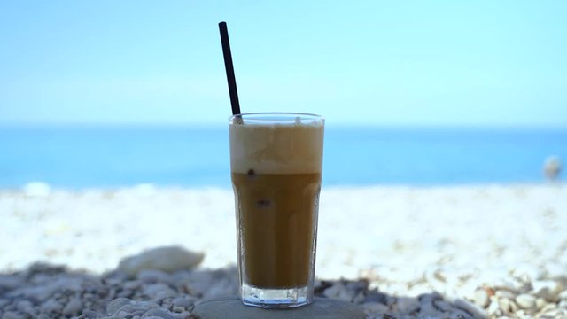Glass of frappe on beach with pebbles and blurry sea background, hand takes the coffee