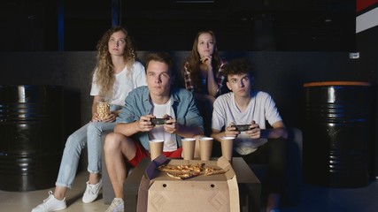 Young friends playing on the game console. Two men holds controllers and plays while girlfriends watches