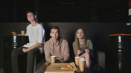 Cheerful company plays in the console game, girl against boy. Eats pizza