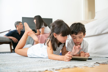Little brother and sister lying on floor in living room and using digital gadgets with learning apps while parents sitting together in background. Internet communication or digital technology concept
