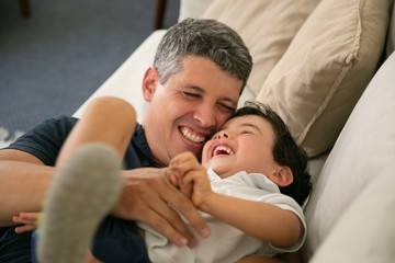 Laughing father tickling his adorable little boy on sofa. Happy attractive Caucasian dad hugging or embracing his lovely son, smiling and playing with child. Fatherhood, leisure and parenthood concept