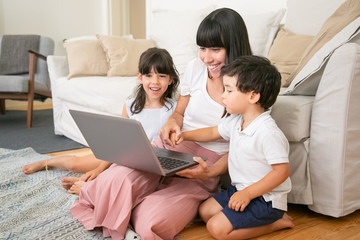 Fototapeta premium Mom and two kids watching funny movie while sitting on floor in living room, using laptop and laughing. Family and home entertainment concept