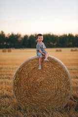 Naklejka na ściany i meble Little boy sitting on a haystack in a field of wheat at sunset