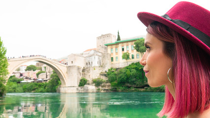 Portrait of a young happy woman with pink hair and a hat who observes the beauty of Mostar and the...