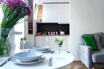 Cozy interior of dining room with small white kitchen and dining table with plate, glass, dinnerware and flower. Modern design and scandinavian style. 