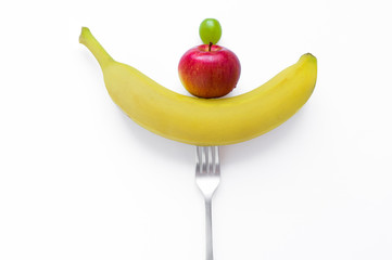 Yellow banana Red apple and green grapes on a fork fresh juicy raw fruits. Vegetarian and vegan concept on white background. Healthy food and diet. Organic vegan health store or store.