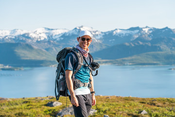 Young Asian man traveller with backpack standing on rock at Senja island in summer season. A man with smiley face enjoying outdoor lifestyle. Norway, Scandinavia