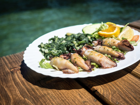 Grilled squid with boiled potatoes and chard (blitva) on white plate with sea on background. Traditional Croatian food. Sunny day.