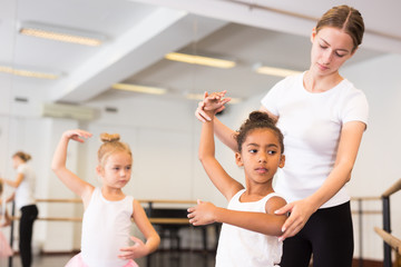 Young female classical dance teacher helping her little girls students in ballet class