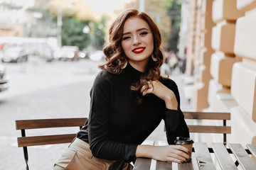 Elegant ginger woman with red lips sitting in outdoor cafe. Stunning curly girl drinking coffee on the street.